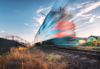 High speed passenger train in motion on the railroad at summer evening. Moving blurred modern commuter train at sunset. Industrial landscape with railway station and blue sky. Transport.  Travel