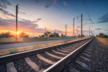Naklejka na ściany i meble Railroad and beautiful sky at sunset in summer. Rural industrial landscape with railway station, blue sky with colorful clouds and orange sunlight, road. Railway platform. Sleepers. Heavy industry.