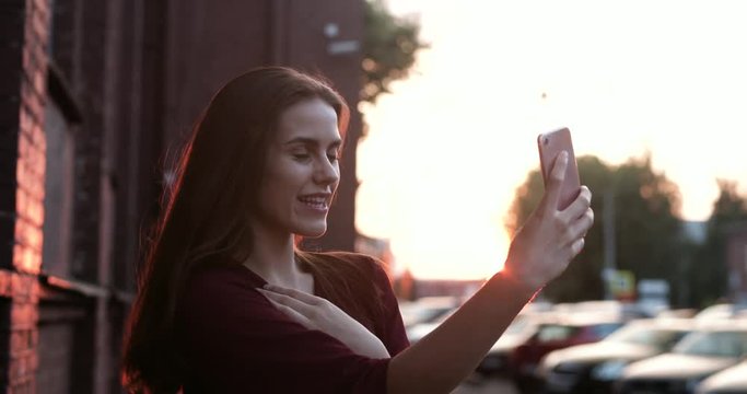 Cute girl smiling and making selfie at sunset. A young girl takes pictures of herself on a smartphone. 