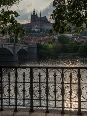Prague castle on summer evening. Back lit seen across Vltava river with a brodge, cast iron railing and sunlit green eaves in foreground