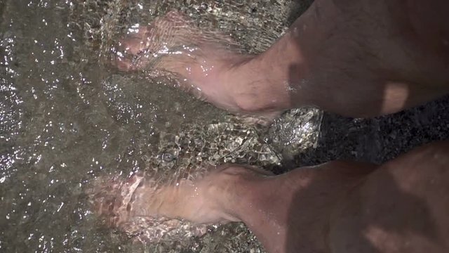 Slow motion wave washes and splashes about man's legs standing on the sand. View from above