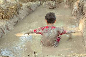 Happy totally dirty boy is sitting and playing in a liquid muddy potter clay improvised basin of mud. Child have fun outdoor. Happy childhood concept