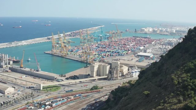 Aerial view of Barcelona industrial harbour with cranes and containers, aside mountain and automobiles moving highway. Drone shot of large cargo port in Barcelona on background of scenic seascape