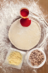 Tomato sauce poured over raw pizza dough on a very floured wooden table. Around it are the ingredients that will be added to the pizza