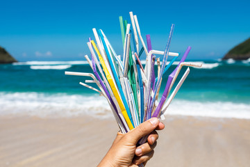 Hand holding heap of used plastic straws on background of clean beach and ocean waves. Plastic...
