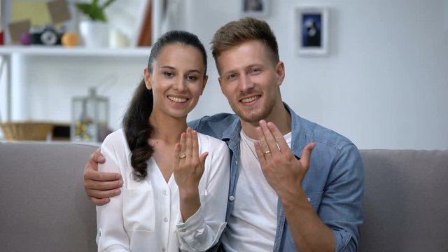 Cheerful couple sitting on sofa, showing hands with engagement rings, love