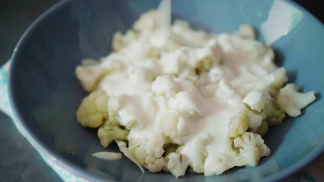 Pouring creamy cheese on cooked cauliflower in a gray plate on dark background, unconventional healthy alternative to Slovak traditional food bryndzove halusky