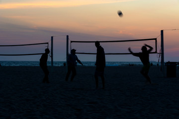 Silhouettes of four volleyball players on the beach at sunset with ocean views and a colorful sky in the background. Huntington Beach, Orange County, California, USA.