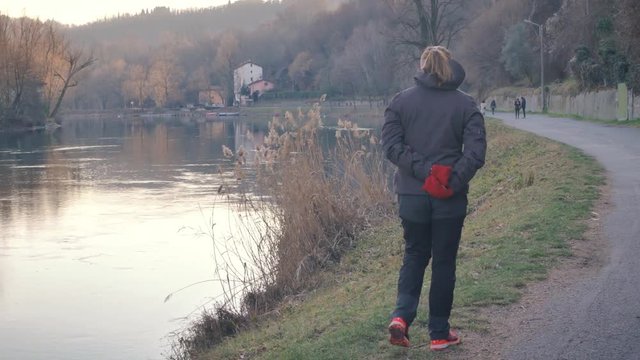 Woman wearing winter jacket and mittens walks beside a river. European countryside in winter. Lady taking a walk outside enjoying nature.