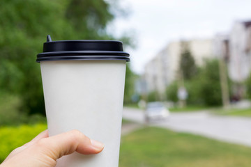 Obraz na płótnie Canvas Hand holding white paper cup with black lid, summer park, car, houses background. Take away, coffee shop concept. Close-up, copy space, mock up