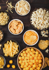 All classic potato snacks with peanuts, popcorn and onion rings and salted pretzels in bowl plates on wooden background. Twirls with sticks and potato chips and crisps with nachos and cheese balls.