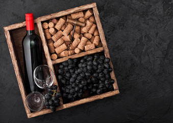 Bottle of red wine and empty glasses with dark grapes with corks and opener inside vintage wooden box on black stone background. Top view