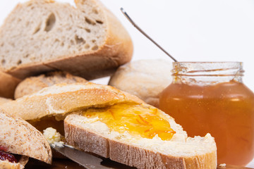 Fresh orange juice, a jar of jam, freshly baked piece of multigrain bread with sunflower seeds, yogurt and more breads in the background
