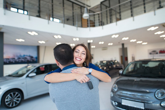 Happy people buying new car at local vehicle dealership showroom. Pretty smiling woman hugging his husband with keys of new automobile.