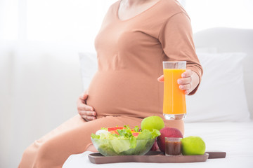 Pregnant woman sitting on bed ,holding  a glass of orange juice.Focus at orange juice glass.