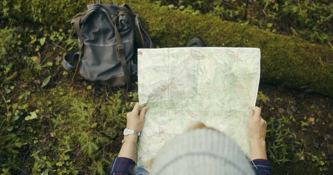 Girl traveler sits on a log in the forest with a map in her hands. See the route on the map. Beautiful place. Top view of the map and the hands of the traveler.