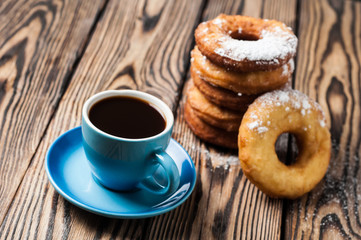 Lot of homemade baked donuts without cream sprinkled of powdered sugar near full cup of hot black coffee lies on old rustic wooden table