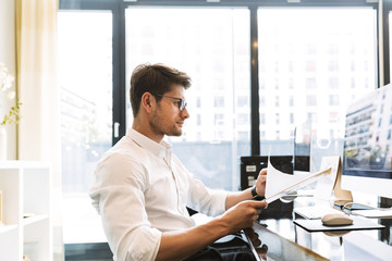 Image of caucasian businesslike man holding clipboard with papers while working on computer in...