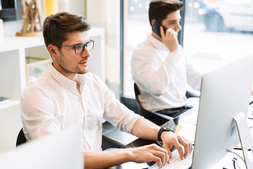 Image of two young men sitting at desk and using smartphone while working on computers in office