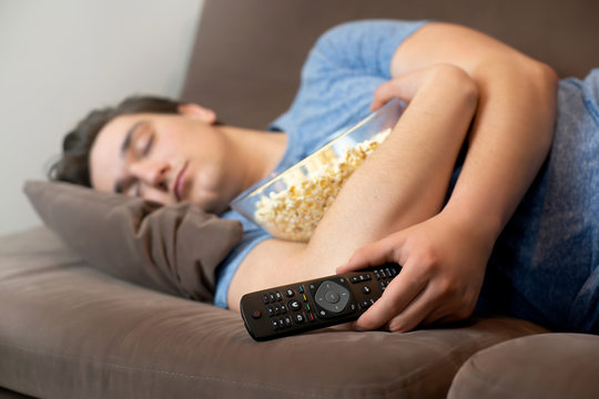 Young Handsome Man Fall Asleep After Watching Thrilling Movie Still Holding Remote Control In One Hand And Popcorn In Other