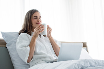 Obraz na płótnie Canvas Young caucasian woman in white pajamas with white holding a cup of coffee and drink and find information from the laptop on a bed.