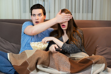 young couple watching movie on the sofa eating popcorn handsome man covering his wife's eyes with his hand both looking frightened
