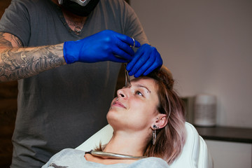 Young Woman getting pierced between her eyes. Man showing a process of piercing with steril medical...