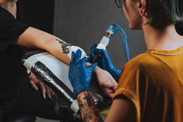 Fototapeta na wymiar Close up tattoo artist demonstrates the process of getting black tattoo with paint. Master works in blue sterile gloves. Master of tattoo fill circuit tattoo.