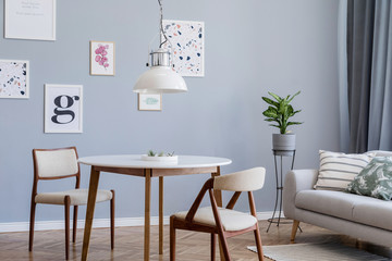 Design scandinavian home interior of open space with stylish chairs, family table  gray sofa, plants elegant accessories and mock up posters gallery wall. Gray background walls. Retro cozy home decor.