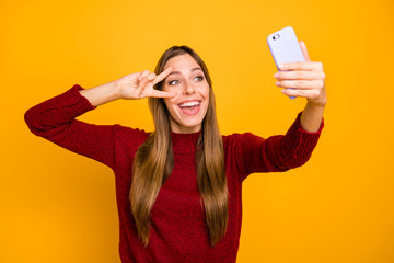 Pretty lady holding telephone hands making selfies showing v-sign wear knitted pullover isolated yellow background