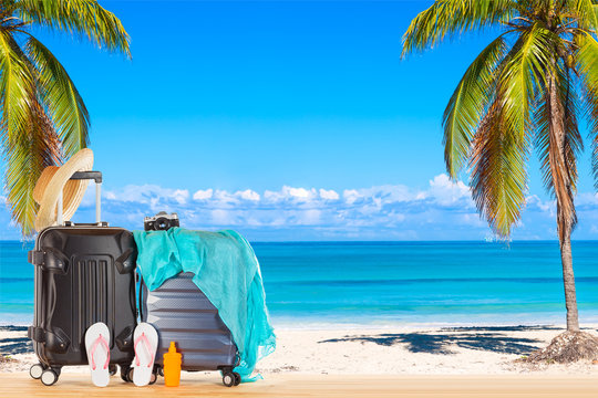 Summer holidays concept. Suitcases baggage with straw hat, blue pareo, flip flops, sunscreen lotion bottle and retro camera against amazing ocean beach with palms and blue sky background.