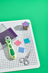 Rotary cutter, ruler, bright square pieces of fabric, stack of bright square fabric pieces, scissors on craft mat, green background