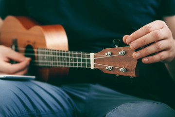 A guy in a blue t-shirt and azure pants adjusts the ukulele using the application in the phone,...