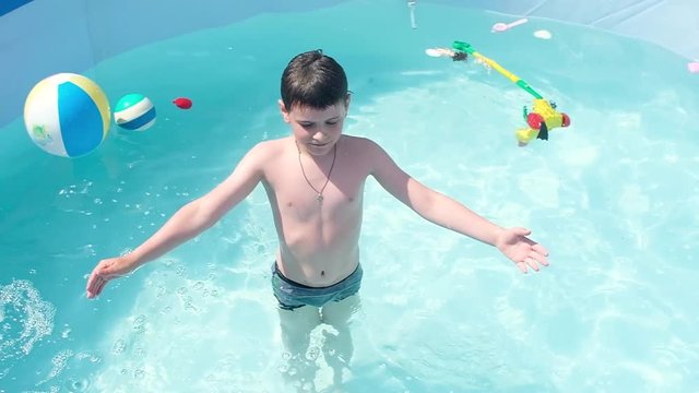 boy with cross on his neck stands in the clear water of the pool with floating toys. Slowly falls back into the water and abruptly jumps out of the water rubbing his eyes. Swimming on hot summer day.