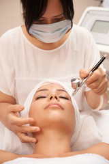 Asian cosmetologist in protective mask doing spa procedure with special equipment for young woman's face while she lying with her eyes closed