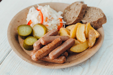 Tasty sausages with fried potatoes, cucumber, cabbage and chlobe in a paper plate on a wooden table.