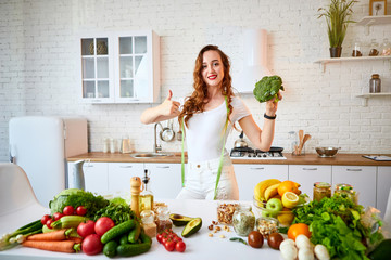 Young happy woman holding broccoli and showing thumbs up in the beautiful kitchen with green fresh ingredients indoors. Healthy food and Dieting concept. Loosing Weight