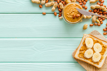 Make sandwiches with banana and peanut butter in glass bowl, knife on mint green wooden background top view copyspace