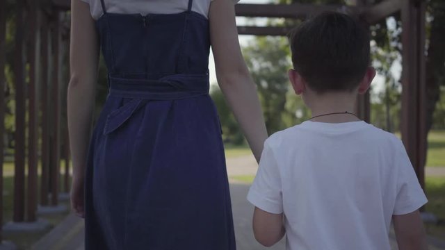 Older Sister Walking With Younger Brother Holding Hands In The Summer Park. Leisure Outdoors. Friendly Relations Between Siblings. Back View