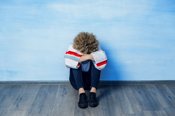 A depressed young guy in a white sweater with a rainbow pattern sits leaning against the blue wall,...