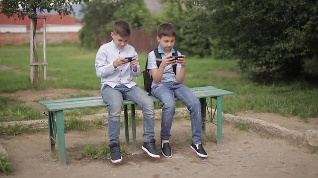 Two boy sitting on the bench and play online games. One boys with backpack. Young boys use their phones