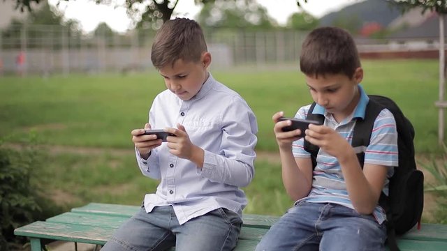 Two boy sitting on the bench and play online games. One boys with backpack. Young boys use their phones