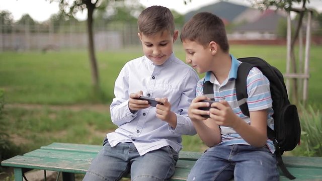 Two boy sitting on the bench and play online games. One boys with backpack. Young boys use their phones