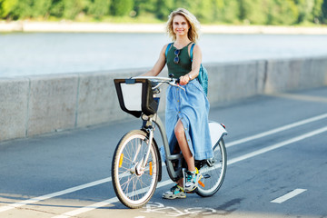 Image of young blonde in long denim skirt sitting on bike on road in city