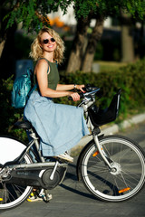 Obraz na płótnie Canvas Photo of young blonde in sunglasses and long denim skirt standing next to bike near green bushes in city