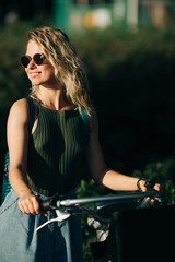 Image of happy curly blonde in sunglasses looking at side in long denim skirt standing on bike next to green bushes in city