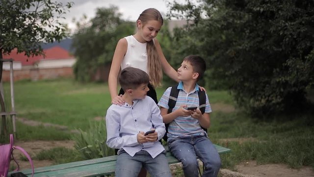 Two boys and girl use their phones during school breack. Cute boys sitting on the bench and play online games. Girl look what boys doing