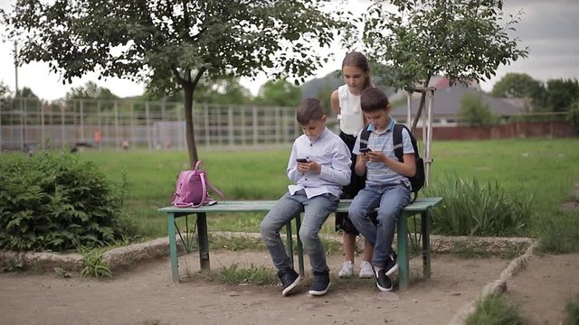 Two boys and girl use their phones during school breack. Cute boys sitting on the bench and play online games. Girl look what boys doing