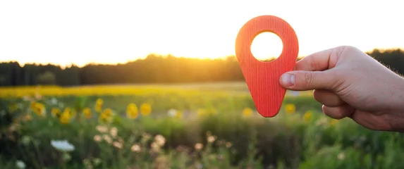 Tuinposter A hand is holding a red location marker in the sunset background. The concept of tourism and travel. Navigation and exploration. Destination. Holiday, vacation. Buying building land. Selective focus © Andrii Yalanskyi