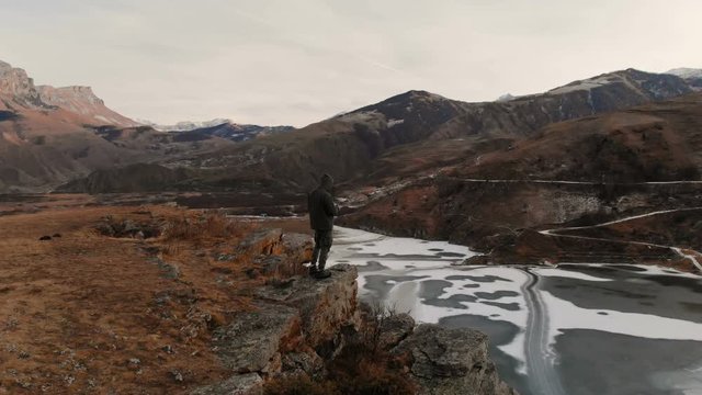 An aerial view of a man in a hood stands on the edge of a cliff near a mountain lake against the backdrop of epic rocks and mountains after sunset at dusk in winter.
